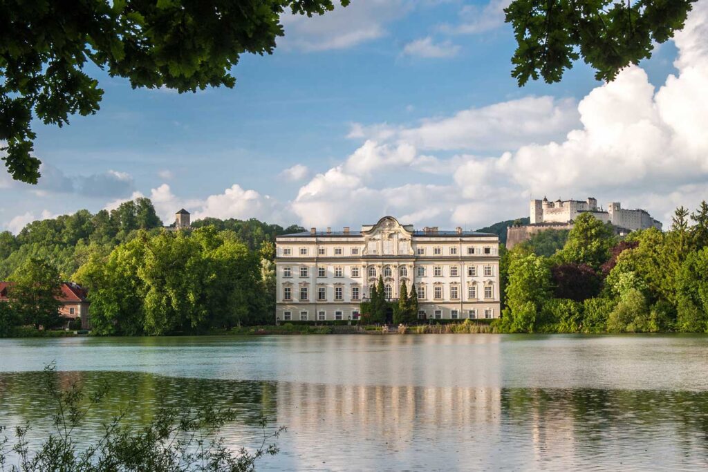 Schloss Leopoldskron majestically overlooks a serene lake, with the imposing fortress Hohensalzburg perched in the distance, a notable stop on the "Private Sound of Maria Tour" in Salzburg.