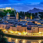 Salzburg by night with its illuminated skyline and the Bavarian Alps in the background.