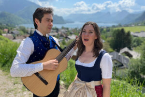 Dana Hammett and Dmitry Iogman, dressed in traditional attire, perform a heartfelt duet against the stunning panorama of the Salzkammergut Lake District, echoing the musical heritage of the region.