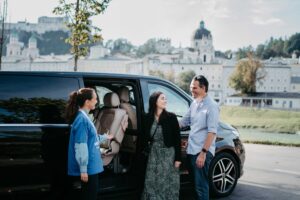 A friendly driver of the sightseeing tour operator weTours Salzburg welcomes a smiling couple next to an elegant black Mercedes Minivan in front of the skyline of Salzburg, ready to embark on a private musical journey through the iconic hills and landmarks that inspired a legendary cinematic story of song and family.