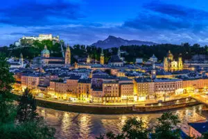 Panoramic view of Salzburg by night from Kapuzinerberg, showcasing the illuminated historic town, the Fortress Hohensalzburg, the Salzburg Cathedral, and the Untersberg in the background, with the Salzach River winding through the scene.